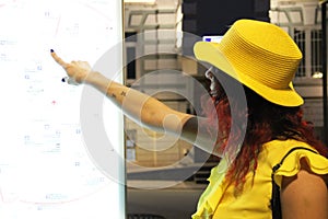 A young woman in a yellow hat looks at the electronic map of the city. A woman in a yellow blouse and hat with her