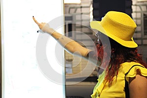 A young woman in a yellow hat looks at an electronic map of the city. A woman in a yellow blouse and hat searches for a