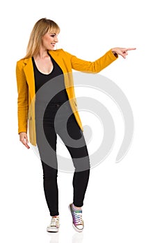 Young Woman In Yellow Jacket Is Pointing To The Side. Full Length, Isolated
