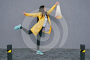 Young woman in a yellow raincoat carrying string bag with fresh bananas and apples on the street against gray wall ,copy