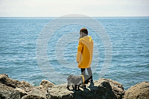 Young woman in yellow raincoat with her dog on background of blue sea water
