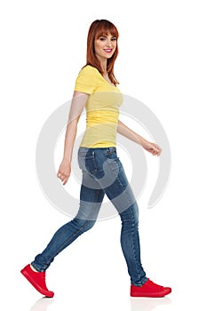 Young Woman In Yellow Shirt, Jeans And Red Sneakers Is Walking And Looking At Camera