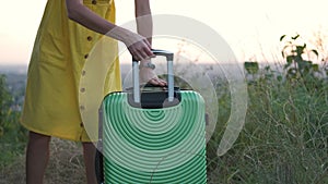 Young woman in yellow summer dress taking green suitcase from car trunk. Travel and vacations concept.