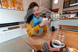 Young Woman in Yellow Sweater And Blue Scarf Drinking Hot Tea at Modern Cafe And Looking at Boyfriend. First Person View