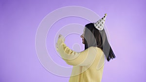 Young woman in yellow sweater with party hat is dancing on a purple background