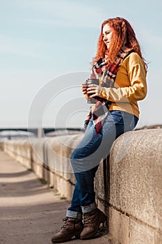 Young woman in yellow sweater sits on embankment and drinks coffee from reusable mug. She is enjoying autumn sunny day.