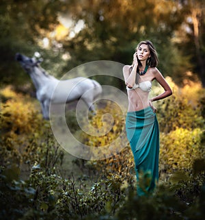 Young women in a blue long skirt and white bra at sunset in forest with a white horse