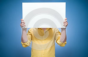 Young women covering her face with white board over blue background, dresses in yellow shirt. Promoting guy showing blank empty