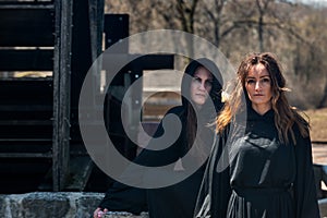 Young Women with dark long hair in black robes in front of an Old Wooden Water Mill. Witches. Halloween concept. Witchcraft