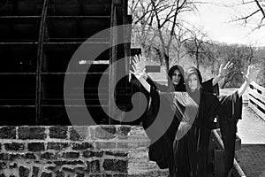 Young Women with dark long hair in black robes in front of an Old Wooden Water Mill. Witches. Halloween concept. Witchcraft