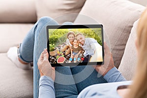 Young woman having remote conversation with happy family on picnic, using tablet computer at home