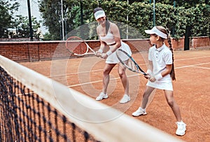 Young woman with llittle girl play tennis standing together by one side on tennis court photo