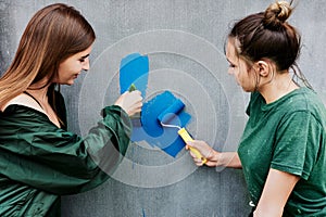 Young women painting gray wall with blue paint using roller and brush.