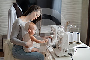 Young woman sews at home and holds a small child. Mom teaches her little son to sew on a sewing machine. Self-isolation