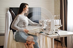 Young woman sews at home and holds a small child. Mom teaches her little son to sew on a sewing machine