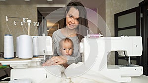 Young woman sews at home and holds a small child. Mom teaches her little son to sew on a sewing machine