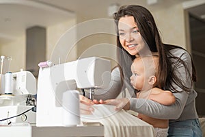 Young woman sews at home and holds a small child. Mom teaches her little son to sew on a sewing machine