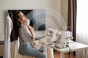 Young woman sews at home and holds a small child. Mom teaches her little son to sew on a sewing machine