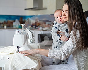 A young woman sews in the kitchen and holds a small child. Mom teaches her little son to sew on a sewing machine. Self