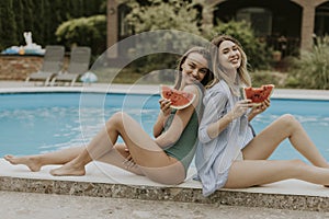 Young women sitting by the swimming pool and eating watermellon in the house backyard