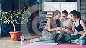 Young women are socialising watching smartphone in gym. They are touching screen, gesturing, chatting emotionally and