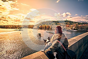 Young women tourist with a puppy dog and a backpack looking at the tourist boat and swans sailing on Vltava river