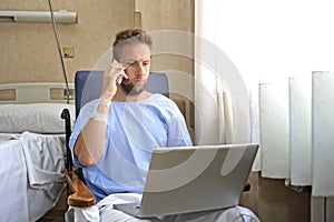 Young workaholic business man in hospital room sick and injured after accident working with mobile phone and computer laptop