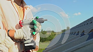 A young worker in a black hat screws a self-tapping screw into a metal tile roof using an electric screwdriver. A roofer