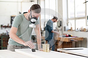 Young worker in a carpenter`s workshop painting wood with brush and respirator