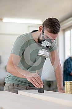 Young worker in a carpenter`s workshop painting wood with brush and respirator