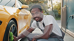 Young worker of self car wash service African bearded man cleaning a wheel, car rims of modern luxury yellow sport car