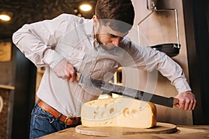 Young worker slice cheese maasdam on wooden board in cheese shop. Man use big cheese knife