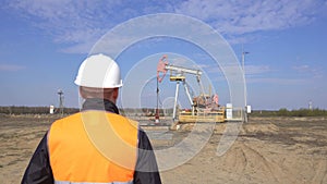 A young working engineer in a signal vest straightens a protective helmet on his head against the background of an oil