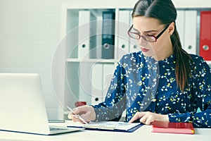 Young woman using laptop and reading annual report document at work. Business woman working at her desk.