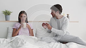 Young worried husband bringing cup of tea to sick wife and supporting her, woman with fever lying in bed, slow motion