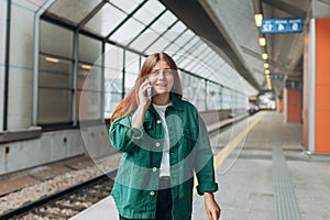 Young worried redhead woman waiting train on station platform, she lost or late, unhappy girl talking on smart phone