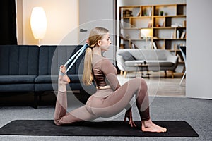 Young yogi woman practicing yoga concept, doing One Legged King Pigeon pose with elastic band, wearing sportswear bra and pants on