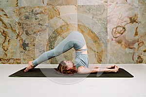 Young yogini practicing Halasana plow pose in pastel blue leggins and top