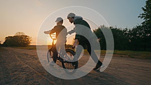 The younger brother learns to ride a bicycle at sunset with the older brother.