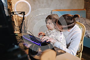 Younger sister sits on brothers knees and put finger on keyboard with neon light. Little girl help brother work on