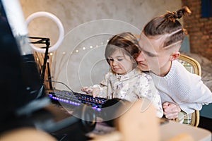 Younger sister sits on brothers knees and put finger on keyboard with neon light. Little girl help brother work on