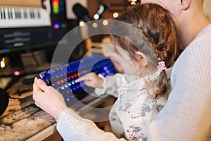 Younger sister sits on brothers knees and put finger on keyboard with neon light. Little girl help brother work on