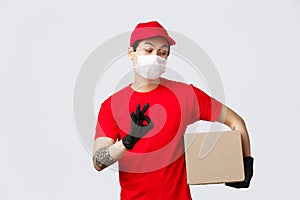 Your parcel is safe. Relaxed asian courier in medical mask and gloves, holding box package and show okay sign, guarantee