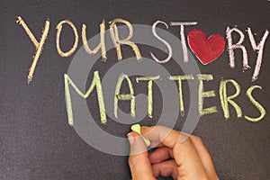 Your story matters phrase handwritten on blackboard with heart symbol instead of O, hand holding green chalk