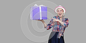 It is yours? Happy beautiful modern grandma in white hat and in checkered shirt standing, surprised holding big gift box with