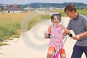 Youth brother teaching his younger sister to ride a bike. Little girl in a pink protective helmet on a pink bike rides