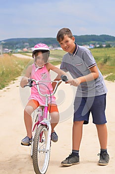 Youth brother teaching his younger sister to ride a bike. Little girl in a pink protective helmet on a pink bike rides