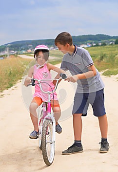 Youth brother teaching his younger sister to ride a bike. Little girl in a pink protective helmet on a pink bike rides