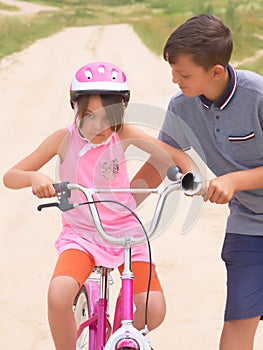 Youth brother teaching his younger sister to ride a bike. Little girl in a pink protective helmet on a pink bike rides