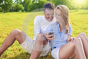 Youth Lifestyle Concept: Laughing Caucasian Couple Sitting Together on the Grass Outdoors with palmtop Player.